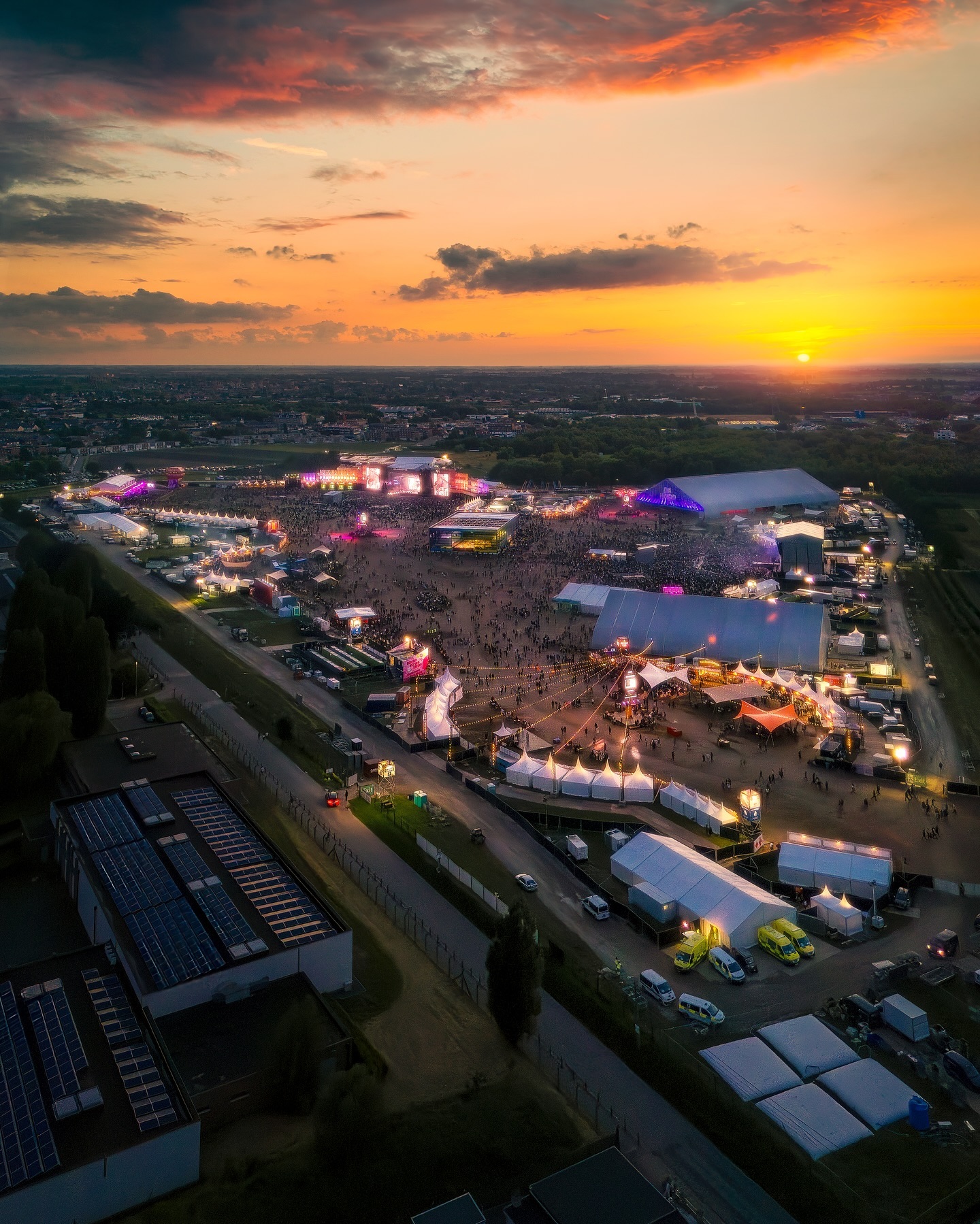Aerial view of a large outdoor festival at sunset with stages, tents, and crowds sprawling across an open field.